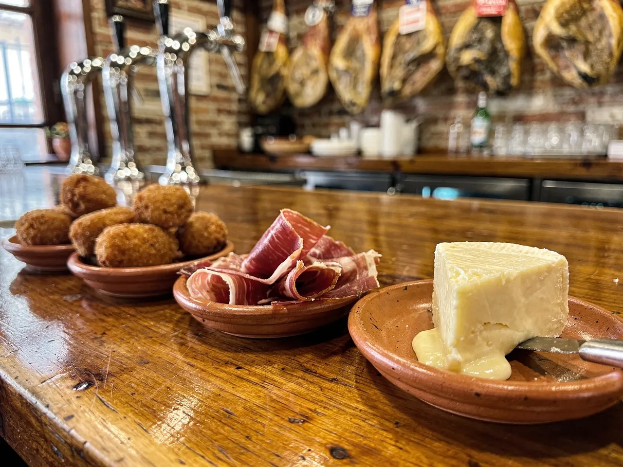 Croquetas, jamón y torta sobre la barra de madera, con jamones colgados al fondo
