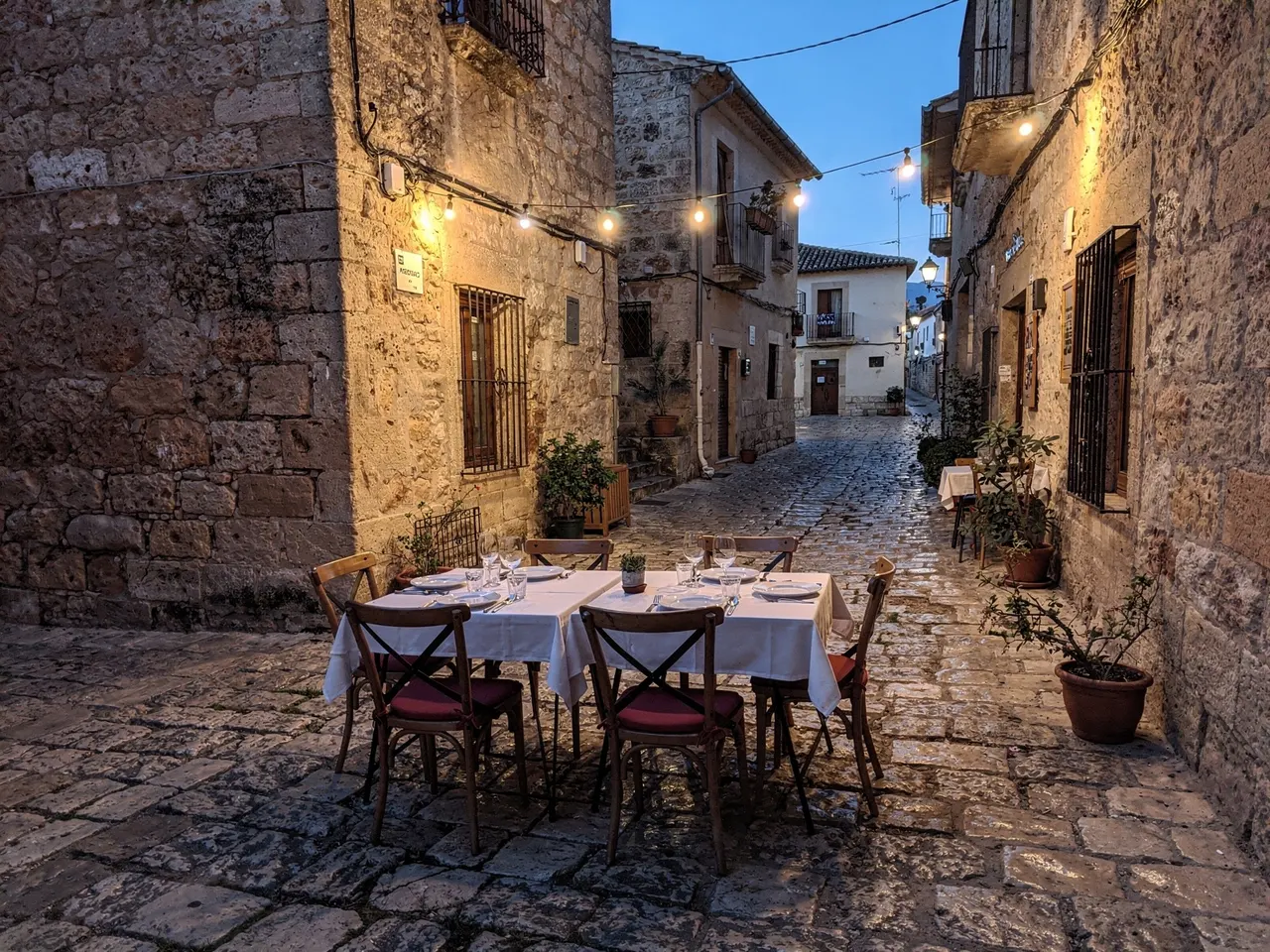 Mesa montada en la terraza de la taberna, en una calle de piedra con guirnaldas encendidas a la hora azul